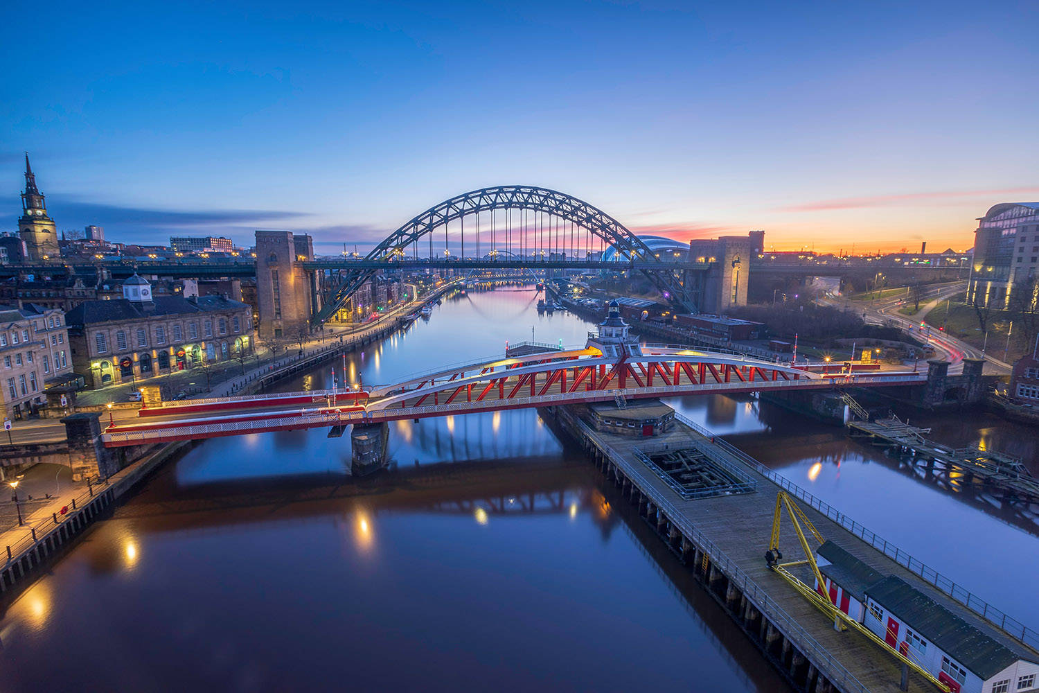 A picture of a bridge over a river in Newcastle. The city is on both sides of the bridge and the sun is just coming up in the horizon.